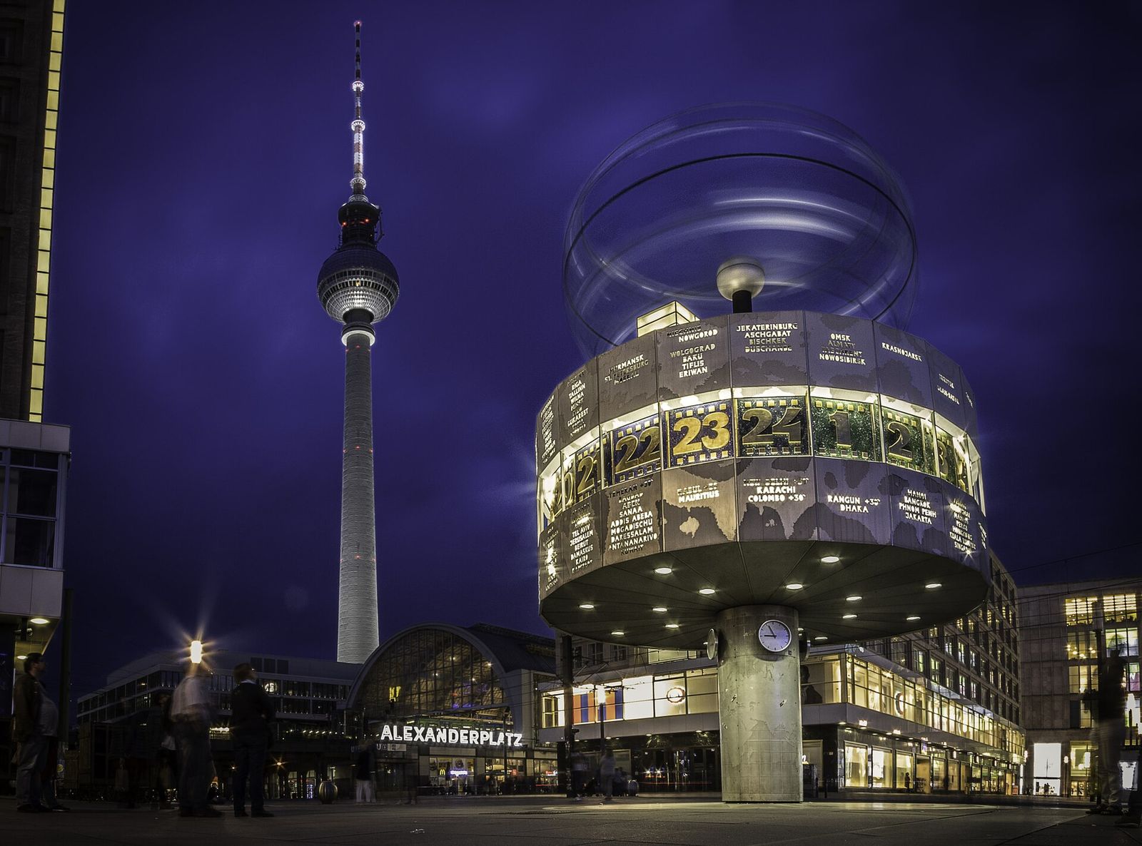 Die beleuchtete Weltzeituhr auf dem Alexanderplatz bei Abendlicht, dahinter Fernsehturm und Bahnhof Alexanderplatz